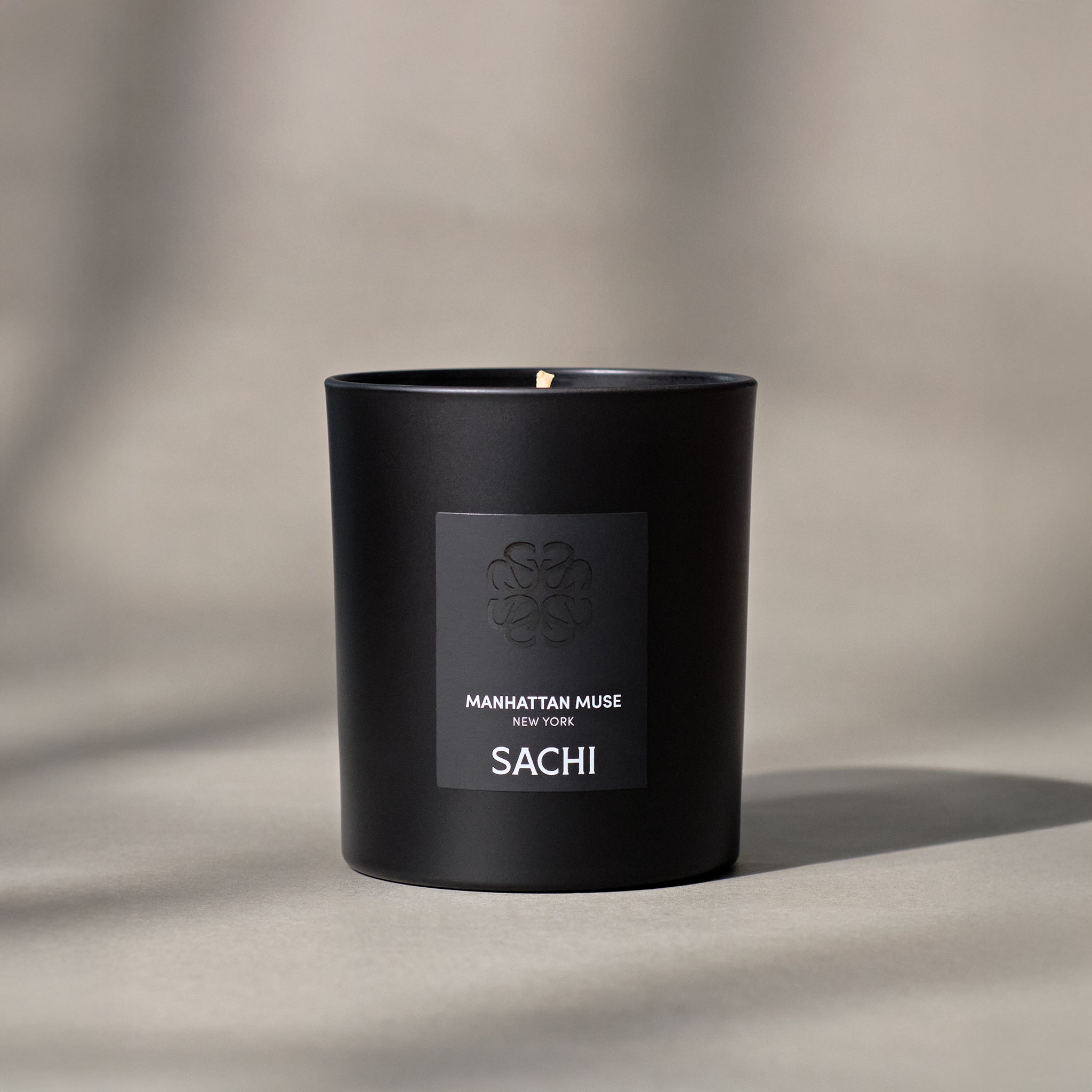 A black candle with the label 'MANHATTAN MUSE' and 'SACHI' in white text, placed on a textured surface with a shadow cast over it.