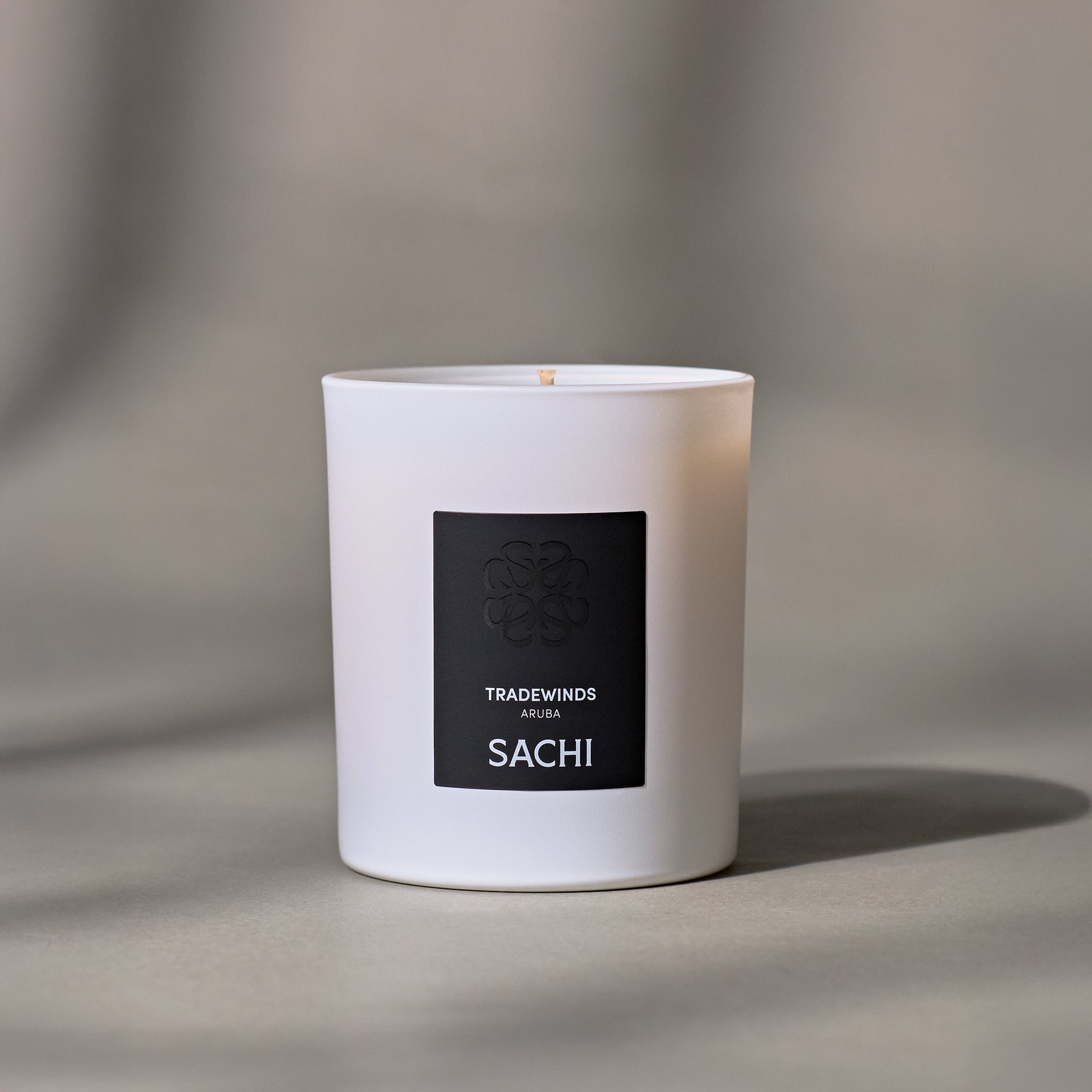 A white candle with a black label that reads 'TRADEWINDS and SACHI', set against a neutral background.